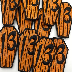 May include: A collection of orange and black coffin-shaped patches. Each patch features the number "13" in black, with a textured, wood-grain appearance. These patches are likely for clothing or accessories.