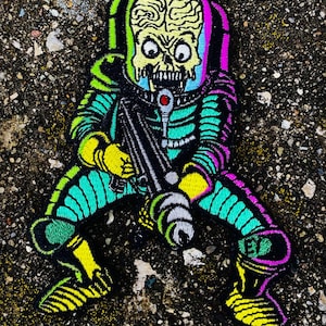 Può includere: Una toppa ricamata colorata che rappresenta un alieno verde con un cervello grande, che indossa una tuta spaziale e tiene una pistola. L'alieno è in posizione accovacciata e ha un'espressione minacciosa.