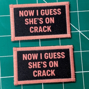 May include: Two rectangular patches with the text "NOW I GUESS SHE'S ON CRACK" in pink lettering on a black background. The patches have a pink border. The patches are on a green cutting mat.