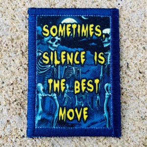 Può includere: Una toppa rettangolare blu con la scritta "SOMETIMES SILENCE IS THE BEST MOVE" in giallo, con uno sfondo blu con scheletri. La toppa ha un bordo blu.