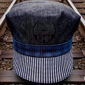 Op de afbeelding: Een denim-stijl treinmachinistpet met een blauw-wit gestreepte rand. De pet heeft een donkerblauwe band en een cirkelvormig embleem met de letters "EMH" en een treinillustratie.