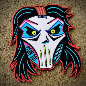 May include: Embroidered patch featuring a stylized skull with long black hair and red accents. The skull has blue and pink details around the eyes and mouth, with yellow eyes. The text "PAIN 101" is visible on the left side of the skull.