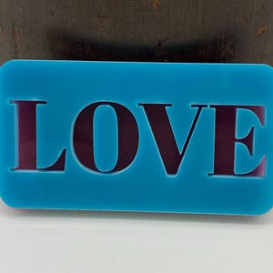 May include: A rectangular bright turquoise plaque featuring the word LOVE cut out in a bold serif font. The letters are filled with a dark, reflective purple material that contrasts with the smooth, matte finish of the surrounding blue surface.