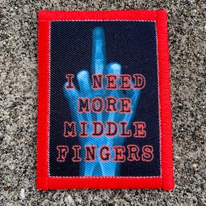 Può includere: Una toppa rettangolare con bordo rosso. La toppa presenta il testo "I NEED MORE MIDDLE FINGERS" in rosso, su un'immagine di mano stilizzata blu su sfondo nero. La toppa è probabilmente da stirare o cucire.