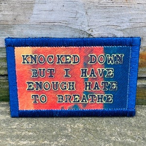 May include: A rectangular patch with a blue border and the text "KNOCKED DOWN BUT I HAVE ENOUGH HATE TO BREATHE" in dark blue. The background has orange, red, and blue tie-dye colors.