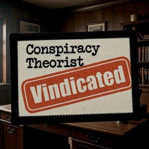 May include: A rectangular patch with the words "Conspiracy Theorist" above a red and white "Vindicated" stamp. The patch has a black border and is set against a blurred background of a wooden desk and bookshelf.