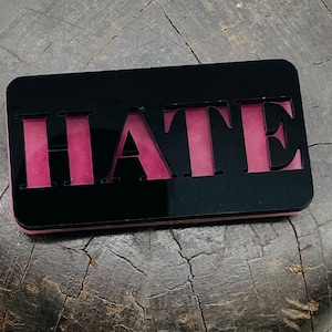 Puede incluir: Un broche rectangular de acrílico negro con la palabra HATE recortada en una fuente serif negrita. Las letras recortadas revelan una capa sólida de color rosa vibrante debajo de la superficie negra, creando un aspecto superpuesto.