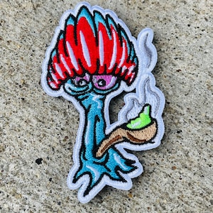 May include: An embroidered patch featuring a cartoon character with a large red and white hat, holding a green pipe. The character has one large eye and a goofy expression.