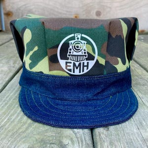 Op de afbeelding: Een camouflage print treinconducteur hoed met een denim rand. De hoed heeft een wit cirkelvormig embleem met een trein illustratie en de initialen "EMH". De hoed is gemaakt van stof en is ontworpen om gedragen te worden.