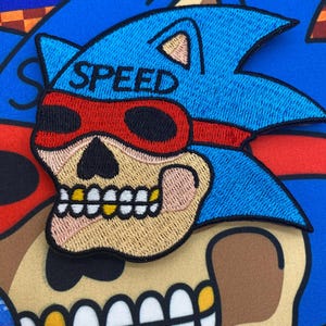以下が含まれることがあります： 「SPEED」と書かれた青いヘッドバンドと赤いサングラスをかけたドクロの青い白の刺繍パッチ