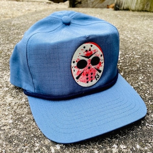 May include: A blue baseball cap featuring a patch with a white mask design, splattered with red paint, and the number 13. The cap has a curved brim and a dark blue rope detail along the base.