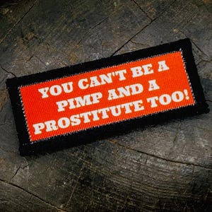 May include: A rectangular embroidered patch with a black border and a bright orange center. The text in white capital letters reads: YOU CAN'T BE A PIMP AND A PROSTITUTE TOO! The patch features a textured fabric surface with visible stitching throughout.