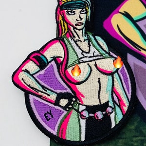 May include: Embroidered patch featuring a stylized female figure with a determined expression. The patch uses vibrant colors, including pink, green, and purple, with flame-shaped accents. The design includes the letters "EY" and is set against a camouflage background.