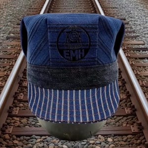 May include: A blue and white striped train conductor hat with a black band and a dark blue top. The hat features a black emblem with a train and the letters "EMH". The hat is displayed on a gray surface.