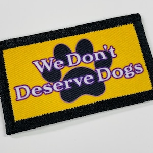 May include: A rectangular patch with a yellow background and a black border. The patch features a black paw print and the text "We Don't Deserve Dogs" in purple and white.