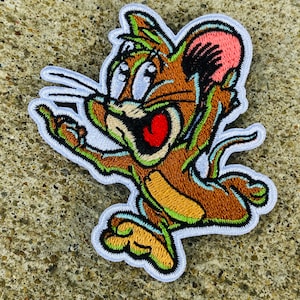 May include: Embroidered patch featuring a cartoon mouse with brown fur, a pink ear, and a red mouth. The mouse is outlined in white and has green and brown accents. The patch is on a textured, gray surface.