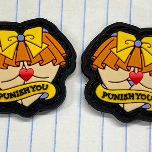 Puede incluir: Dos parches con bordes negros con un diseño de dibujos animados. El diseño presenta una cara con un lazo amarillo, cabello naranja y un corazón rojo. Una pancarta amarilla en la parte inferior dice "PUNISH YOU".