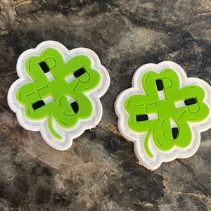 May include: Two four-leaf clover charms featuring a bright green surface with a white border. Each clover has black rectangular cutouts forming the letters P, R, I, and G, with additional small black rectangular accents integrated into the design.