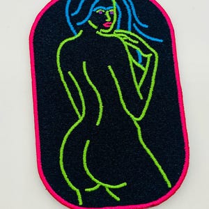 Peut inclure: Un patch brodé noir avec un contour rose. Le patch présente un dessin au trait vert d'une figure nue aux cheveux longs.