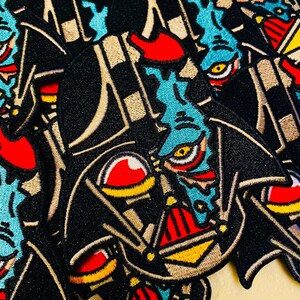 May include: A close-up of a black and gold embroidered patch featuring a blue and red alien character with a large eye. The alien character is wearing a black cape and has a red and yellow helmet. The patch is a popular design for fans of science fiction and fantasy.