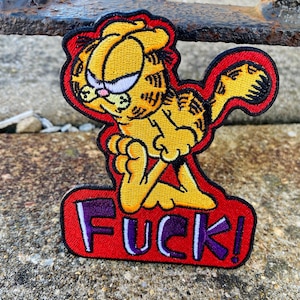 May include: An embroidered patch featuring the cartoon character Garfield in yellow and orange with black stripes, depicted with a grumpy expression. Below the character, the word FUCK! is stitched in purple letters on a red background.