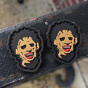 May include: Two black shoe charms featuring a smiling face with a tan complexion, black curly hair, and a red mouth. The charms have a black border and are likely made of rubber or a similar material. The design is a pop culture reference.