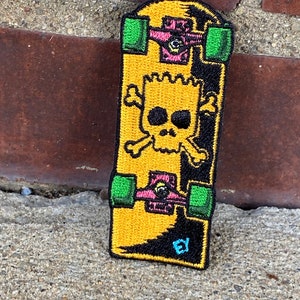 May include: Embroidered patch featuring a yellow skateboard design with a skull and crossbones graphic. The skateboard has green wheels and pink trucks. The patch has black and yellow details, with the initials "EY" in the corner.