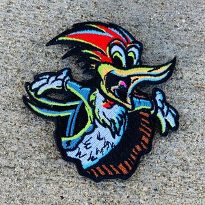May include: Embroidered patch featuring a cartoon bird with a black body, red, yellow, and blue accents. The bird has a long beak and is surrounded by a black border. The patch is likely for clothing or accessories.