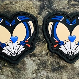 Könnte beinhalten: Zwei schwarze herzförmige Patches mit blauen und weißen Designs. Die Patches zeigen einen stilisierten Schädel mit einem blauen Herzen in der Mitte. Der Schädel ist von einem weißen fächerartigen Design umgeben.