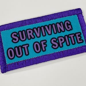 May include: Purple and teal embroidered patch with the text "SURVIVING OUT OF SPITE" in white lettering.