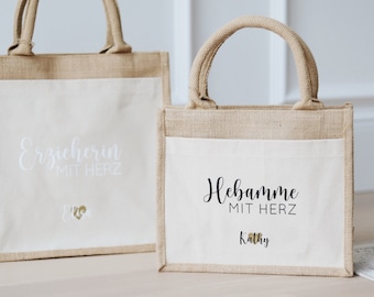 Puede incluir: Dos bolsas de mano beige con inserciones de tela blanca. La bolsa de la izquierda tiene el texto "Erzieherin mit Herz" bordado en blanco. La bolsa de la derecha tiene el texto "Hebamme mit Herz" impreso en negro con el nombre "Kathy" impreso debajo.