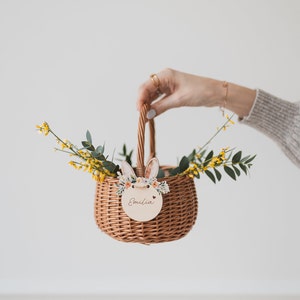 May include: A brown wicker basket with a wooden bunny rabbit cutout and the name "Emilia" on a tag. The basket is filled with yellow flowers and green leaves.