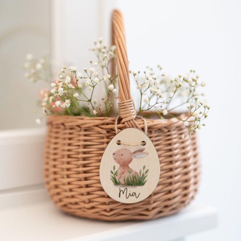 May include: A small wicker basket filled with white baby's breath flowers and a wooden Easter egg tag with a bunny illustration and the name "Mia".