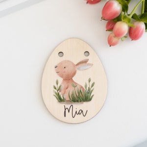 May include: A wooden Easter egg ornament with a brown bunny rabbit illustration and green grass. The egg has two holes at the top and the name "Mia" is written below the bunny.