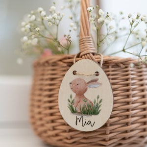 May include: A small wicker basket with a white flower arrangement and a wooden Easter egg tag with a bunny illustration and the name "Mia" written on it.