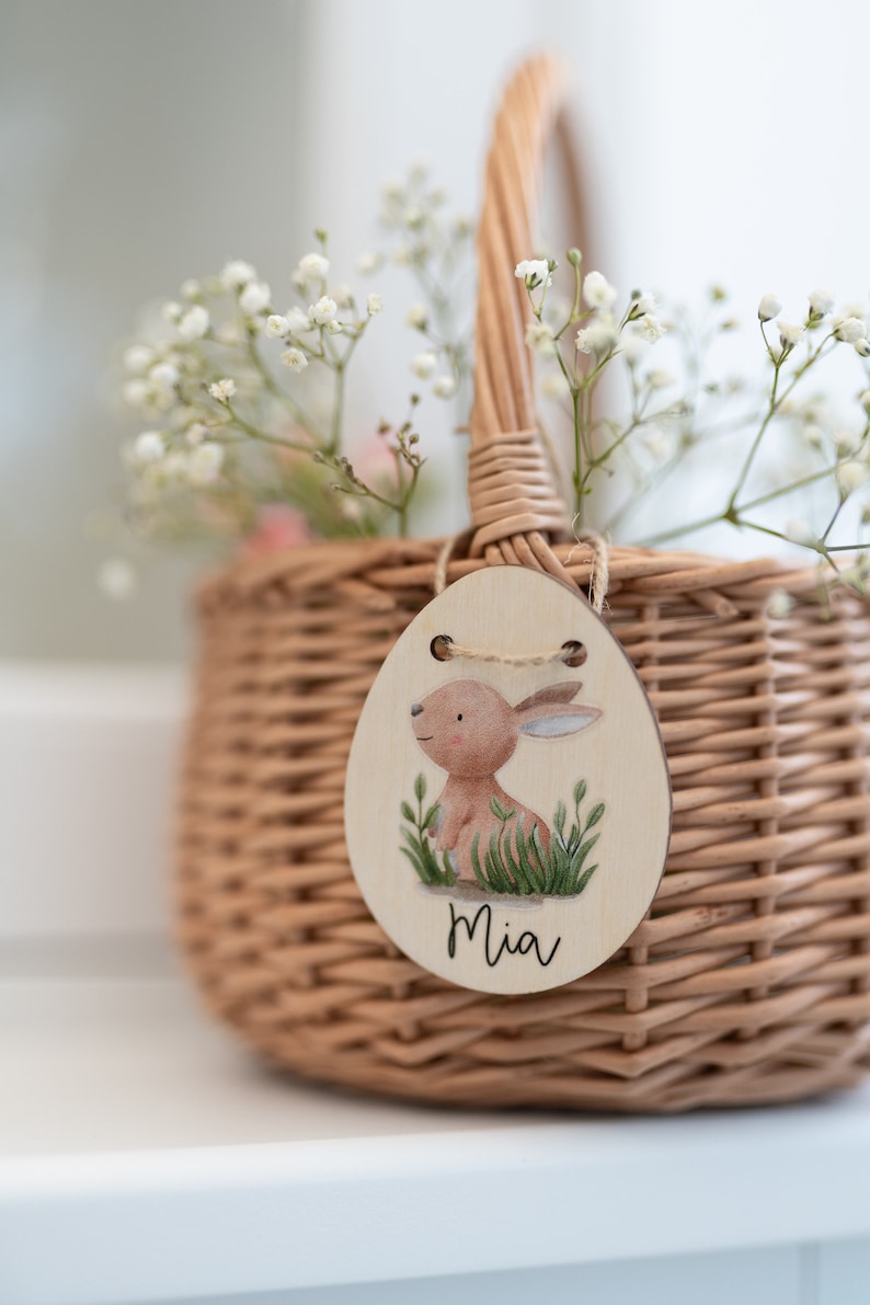 May include: A small wicker basket with a white flower arrangement and a wooden Easter egg tag with a bunny illustration and the name "Mia" written on it.