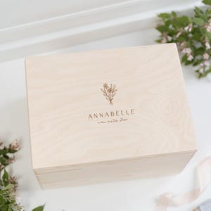 Könnte beinhalten: Eine helle Holzkiste mit einem floralen Design und dem Text "ANNABELLE" und "mein erster jahr" auf dem Deckel.