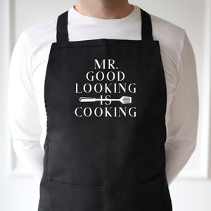 Könnte beinhalten: Schwarze Schürze mit weißem Text, der "Mr. Good Looking Is Cooking" lautet, mit einer Spatel- und Messergrafik.