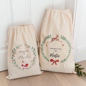 May include: Two white canvas gift bags with drawstring closures. The bags feature a green wreath design with a red bow and the text "Vom Nikolaus fur" followed by a name. One bag has a reindeer illustration and the name "Mateo". The other bag has a Santa Claus illustration and the name "Mila".