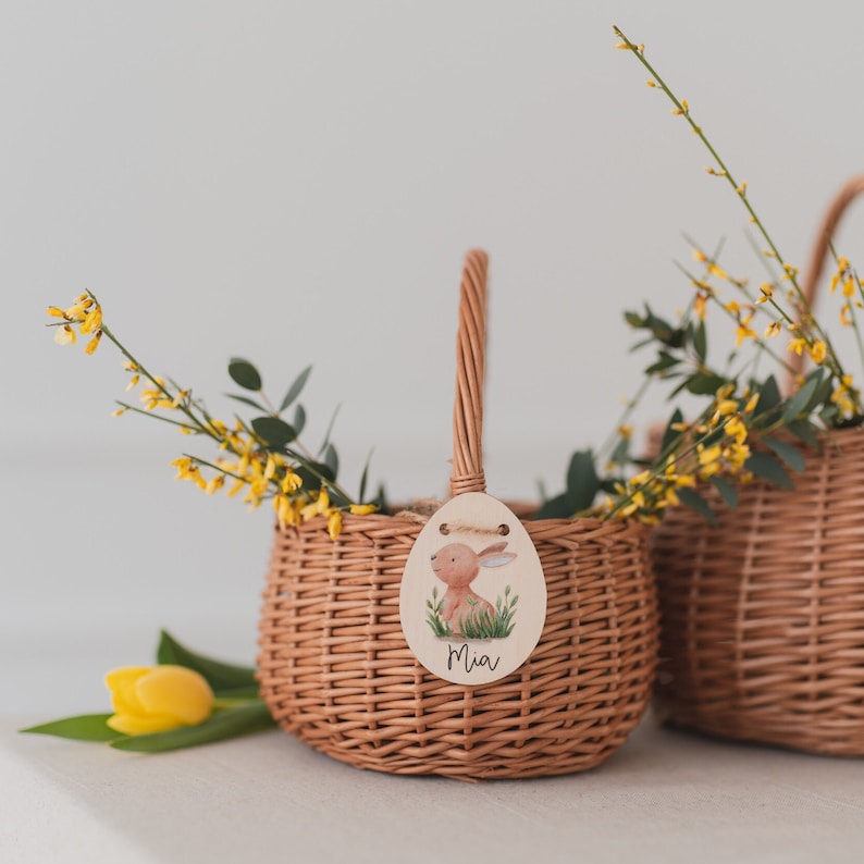 May include: Two brown wicker baskets with handles, one with a wooden tag that says "Mia" and has a cartoon bunny illustration. Both baskets are filled with yellow flowers.