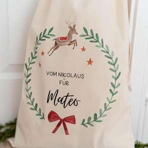 May include: A white canvas gift bag with a red bow, a green wreath, a reindeer with a red saddle, and the text "VOM NIKOLAUS f&uuml;r Mateo".
