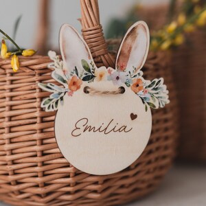 May include: A wooden Easter basket tag with a bunny rabbit design, a floral wreath, and the name "Emilia" written on it.