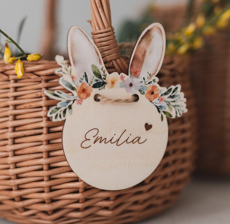 May include: A wooden Easter basket tag with a bunny rabbit design, a floral wreath, and the name "Emilia" written on it.