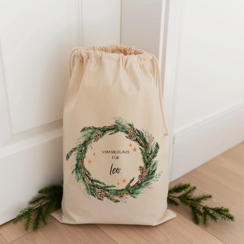 May include: A white canvas sack with a drawstring closure. The sack has a green and brown pine wreath design with gold stars and the text "Vom Nikolaus fur Leo".