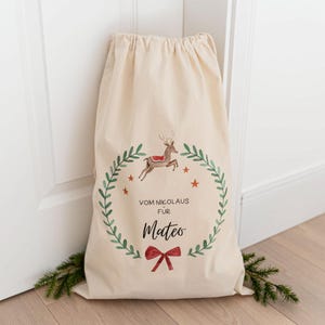 May include: A white canvas sack with a drawstring closure. The sack has a festive design featuring a reindeer, a wreath of green leaves, and red stars. The text "Vom Nikolaus fur Mateo" is printed on the sack.