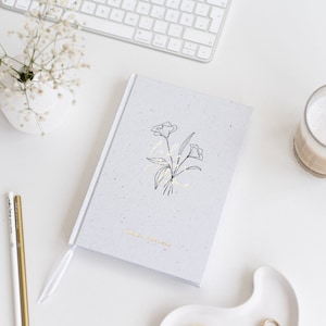 Könnte beinhalten: Ein weißes Journal mit einem grauen, gesprenkelten Einband. Der Einband zeigt eine schwarze Linienzeichnung von Blumen und den Text "Live your life" in goldenen Lettern. Das Journal hat auch den Text "Mini Edition" auf der Unterseite des Einbands.