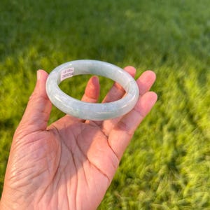 May include: A smooth, translucent pale green jade bangle bracelet with a rounded profile. A small white rectangular sticker is attached to the inner surface of the bangle, featuring the printed text 8626082 700133 in black ink.
