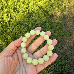 May include: A stretch bracelet composed of smooth, round, pale lime green beads. The beads have a glossy, uniform finish and are strung together in a circular shape, creating a continuous loop of light-colored, spherical gemstone-like elements.
