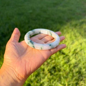 May include: A smooth, circular bangle bracelet crafted from translucent stone with a polished finish. The material features a pale, milky white base color with distinct, irregular streaks and patches of deep forest green throughout.