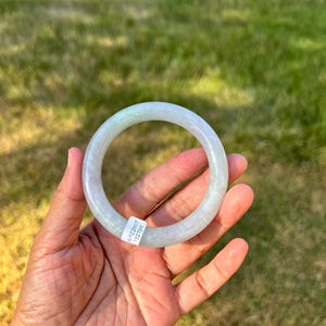 May include: A smooth, circular bangle bracelet crafted from pale, translucent green jadeite stone. The bangle features a polished, rounded surface and includes a small white rectangular sticker with the printed text WPZ2607 152796.